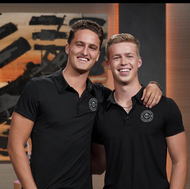 Two young men in matching black Storage Scholars polo shirts smiling together on the Shark Tank set
